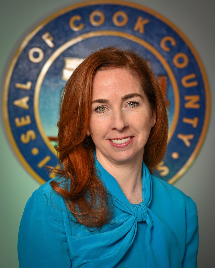 Portrait of a woman with red hair wearing a turquoise blouse, smiling in front of the Cook County seal.