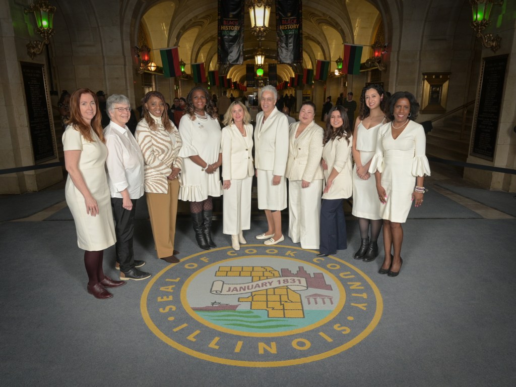 A group of women dressed in white and cream attire standing together in a historic building, with the Cook County seal visible on the floor and decorations for Black History Month in the background.